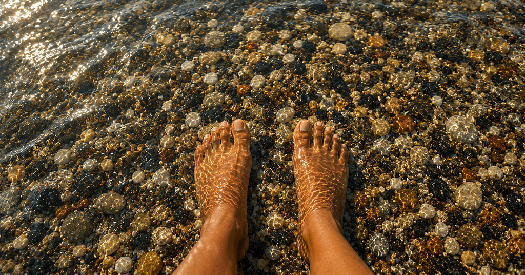 Bare feet with a warm caramel skin tone standing in shallow water over colorful pebbles, symbolizing reflection and the journey of living with flat feet.