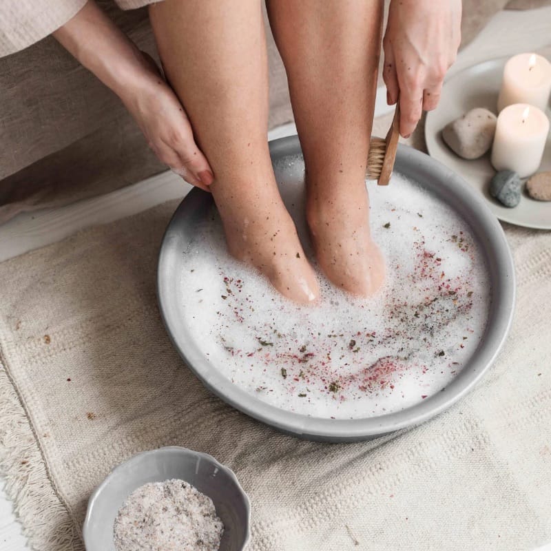 person soaking their feet in warm, foamy water with candles nearby, showing heat therapy for stiff feet and tight muscles.